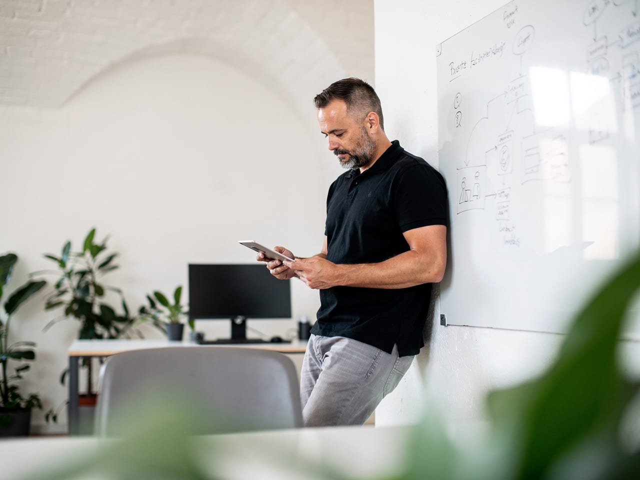 Mitarbeiter lehnt mit einem Tablet an der Wand neben einem Whiteboard